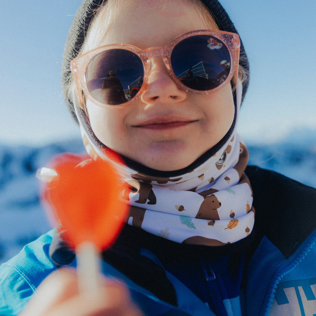Child wearing sunglasses and a patterned Phunkshun Wear necktube, holding an red lollipop with a blurred natural background